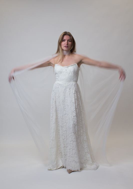 Woman wearing a white lace dress with a tulle bridal cape with a plain background