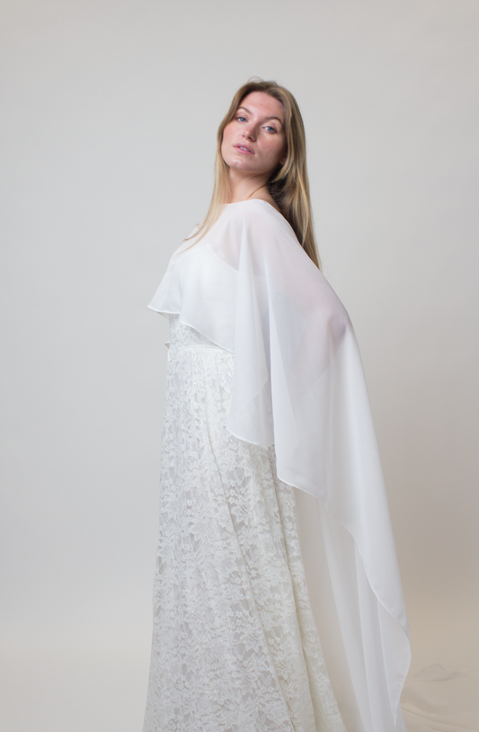 Woman wearing a white lace dress and a chiffon bridal cape against a plain background